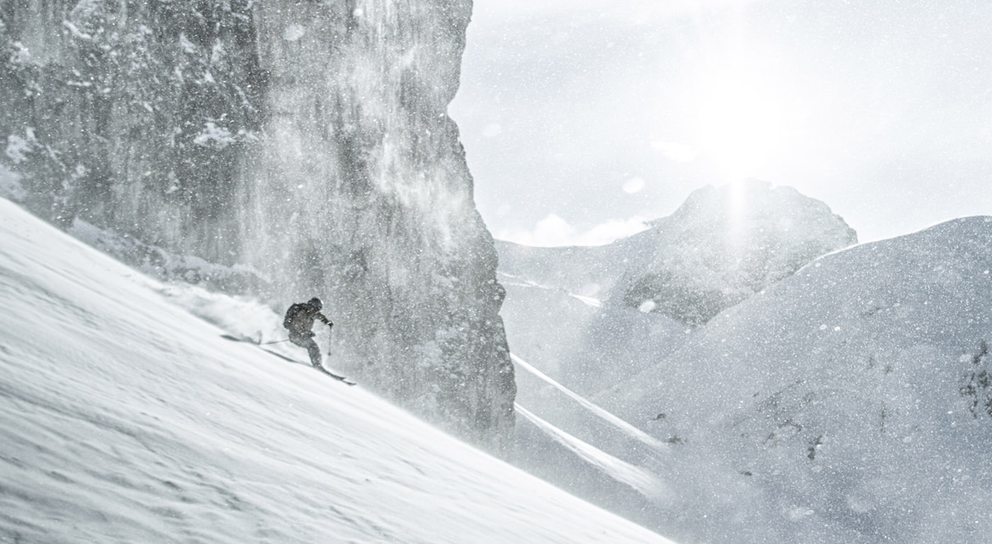 Ein unerschrockener Skifahrer gleitet durch eine Flut von frischem Pulverschnee und fährt einen steilen Berghang hinab, während das Sonnenlicht durch den Schneenebel dringt.